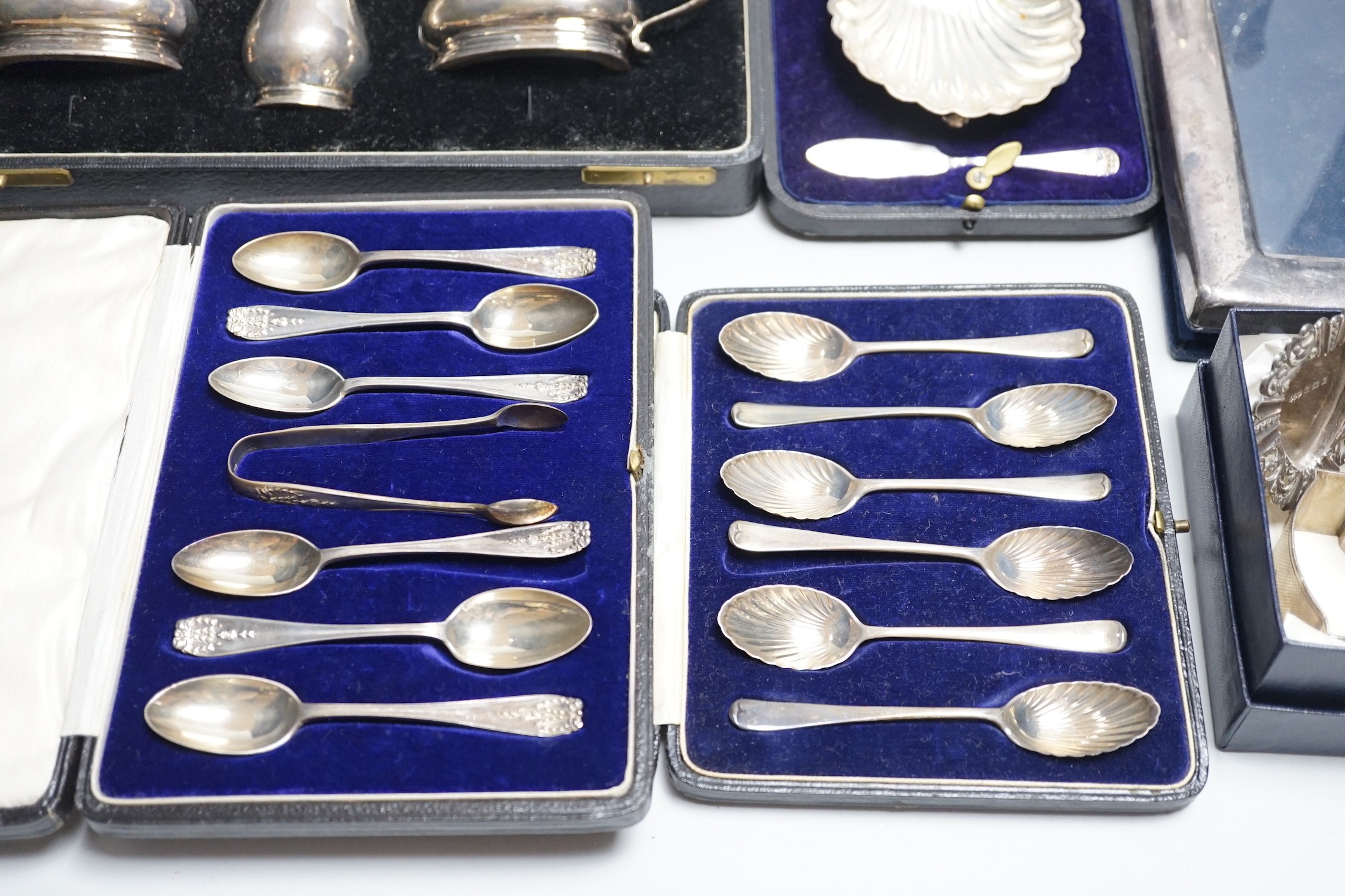 A quantity of mixed silver ware including a pair of modern candlesticks, 15cm, weighted, a cased George V three piece condiment set, cased butter shell and knife, two cased sets of teaspoons, a mounted photograph frame,